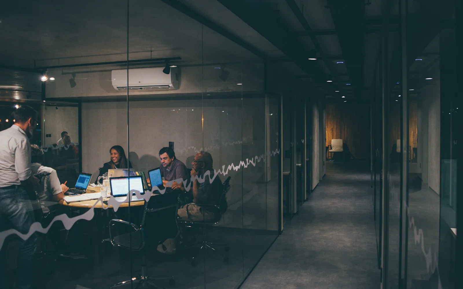 Marketing team meeting around laptops in a glass-walled conference room at night, seen from an office corridor