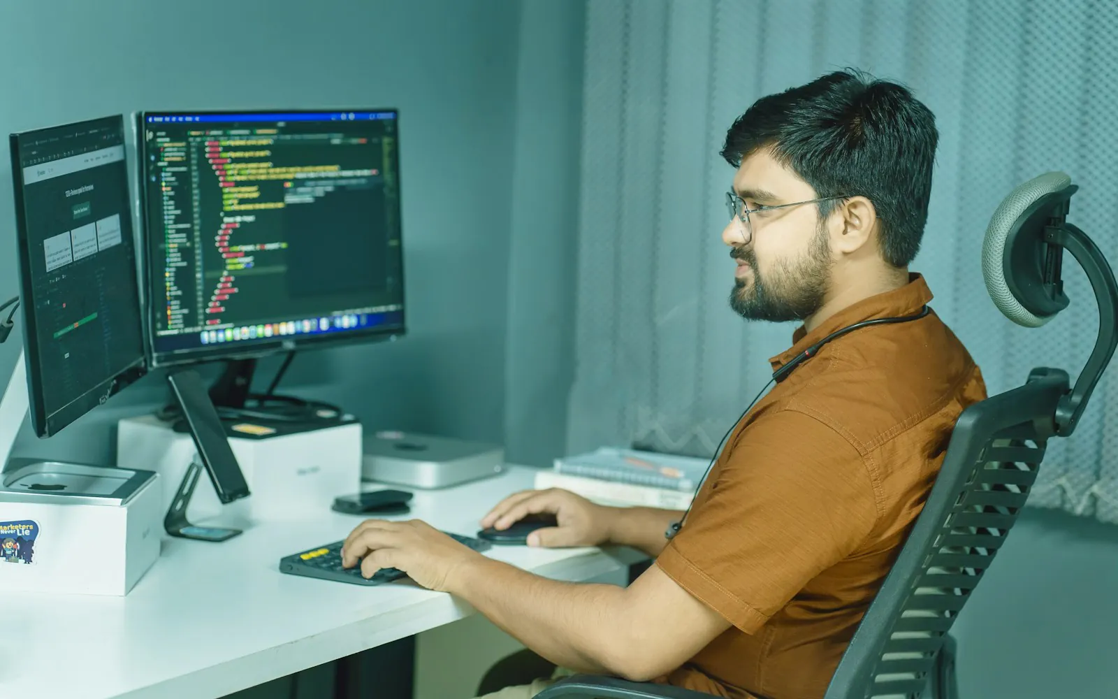 South Asian man in glasses working at a desk with two monitors showing code and a web page, in a softly lit office