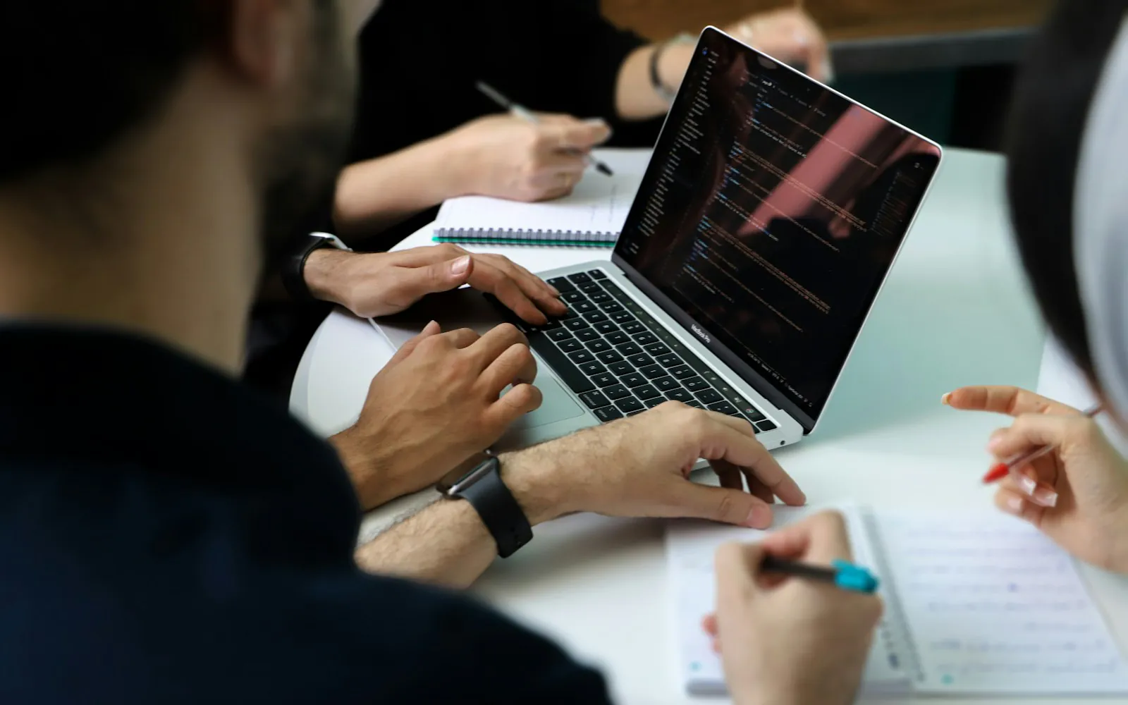 Colleagues around a table with a laptop and notebooks, one typing while others take notes during a planning discussion