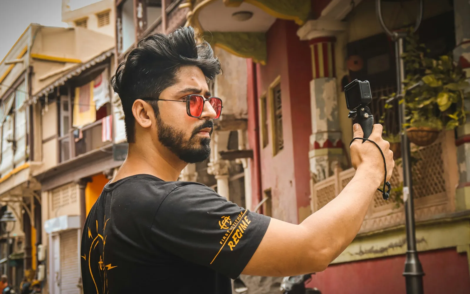 Bearded man in red sunglasses filming himself with an action camera on a handheld grip in an old city street