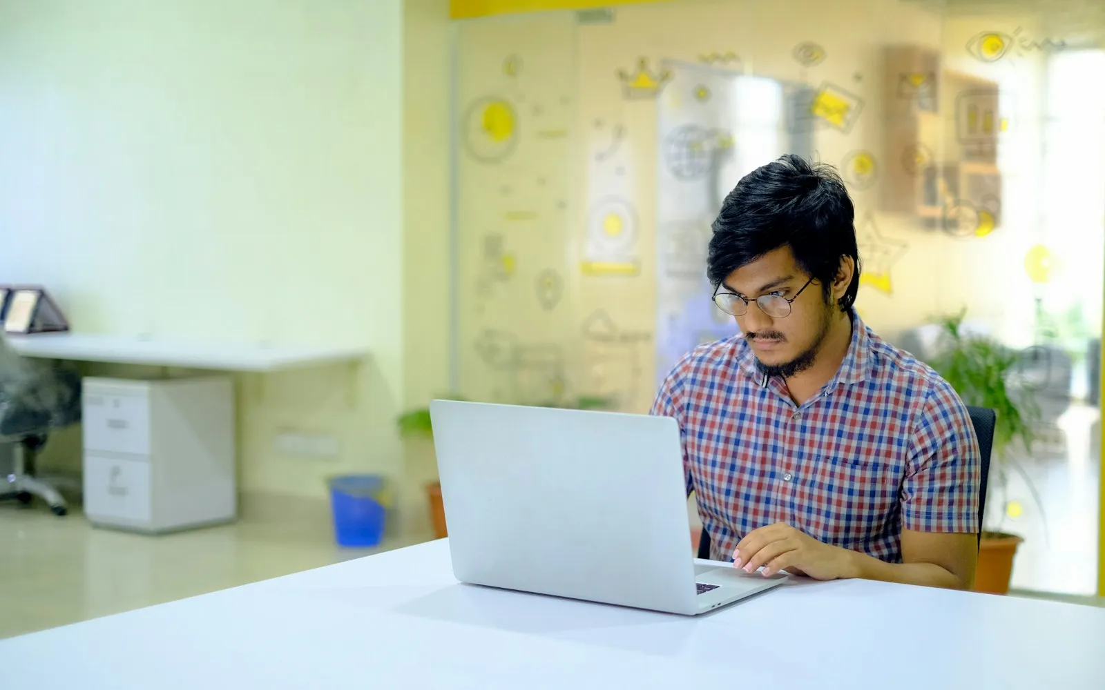 Young man in a checked shirt working on a laptop at a white desk in a bright office