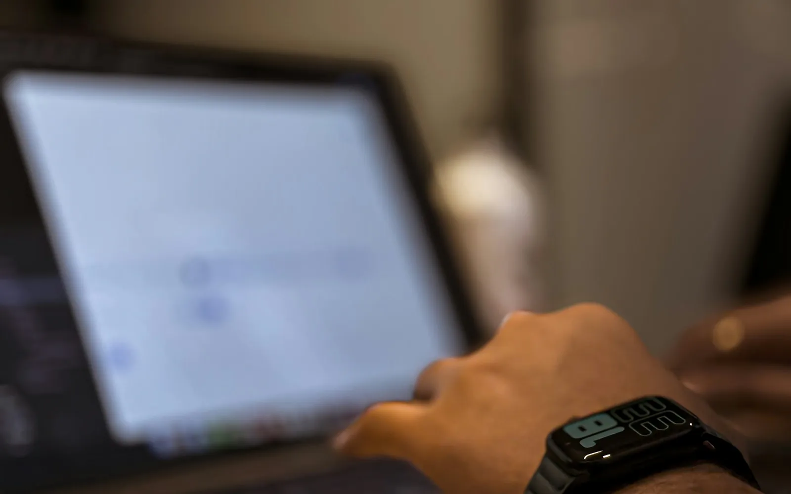 Close-up of a person's hand with a smartwatch reaching toward a laptop trackpad, the screen softly blurred in a dim workspace