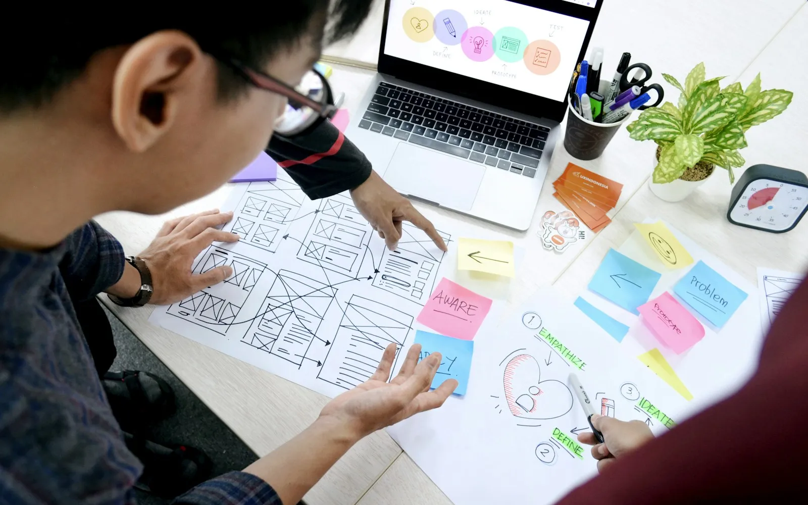 Designers reviewing wireframe sketches and sticky notes beside a laptop showing empathize, define, ideate, prototype, test