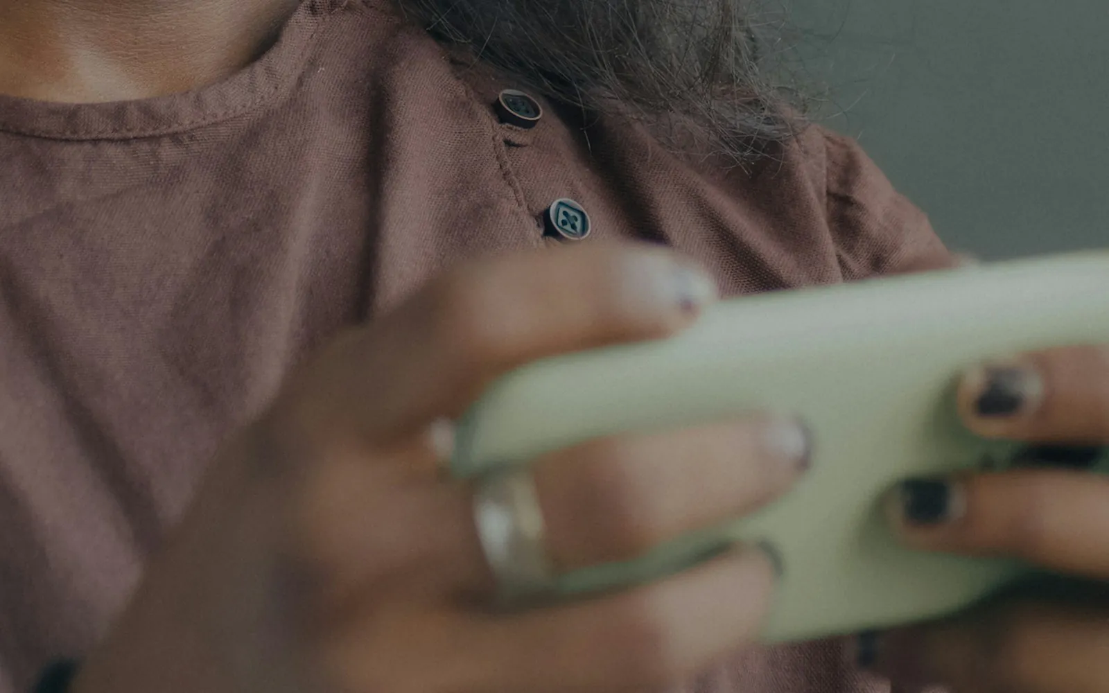 Close-up of a person's hands holding a pale green smartphone in landscape orientation, wearing a ring and a brown linen shirt