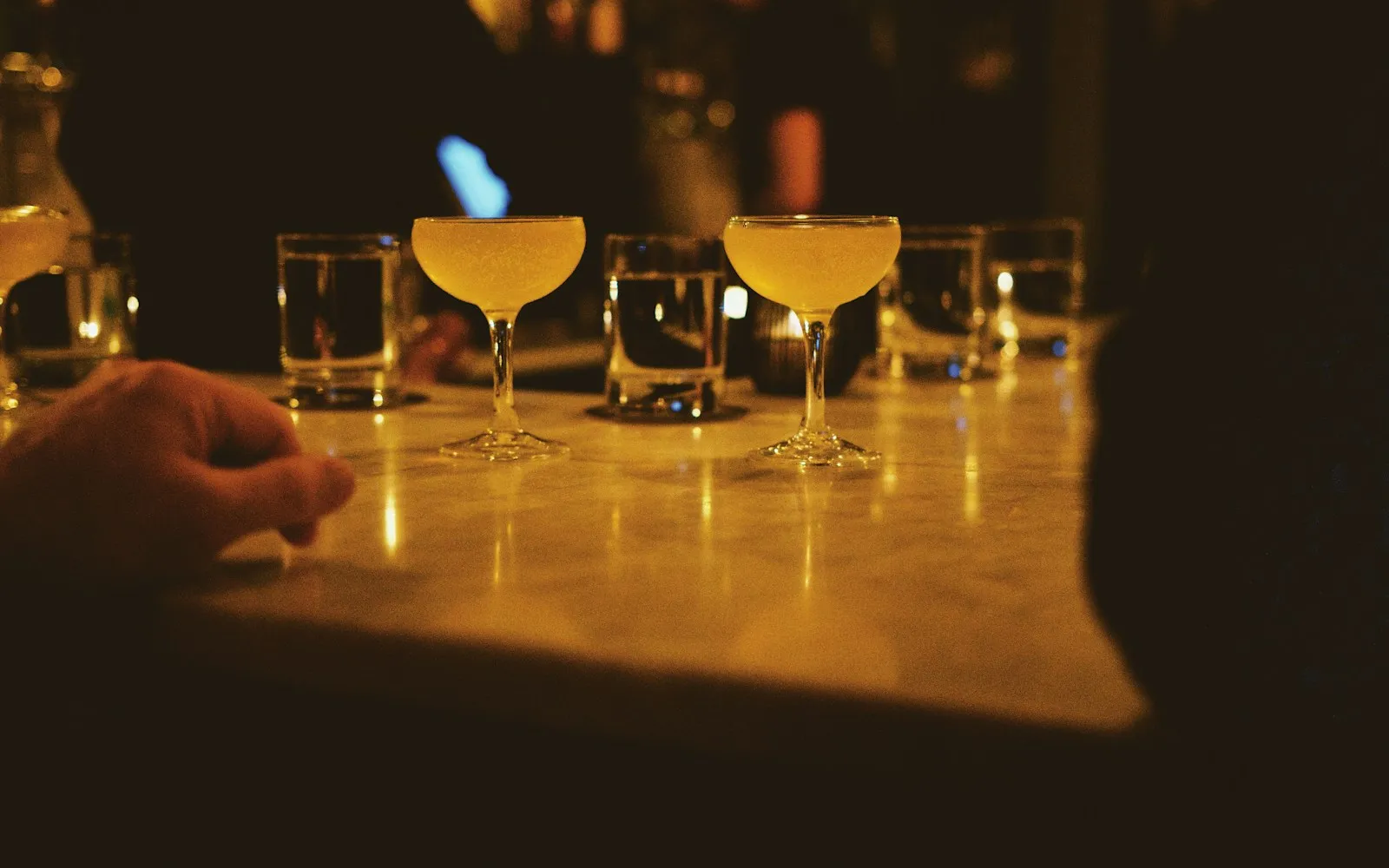 Two cocktails in coupe glasses and water glasses on a dimly lit marble bar counter