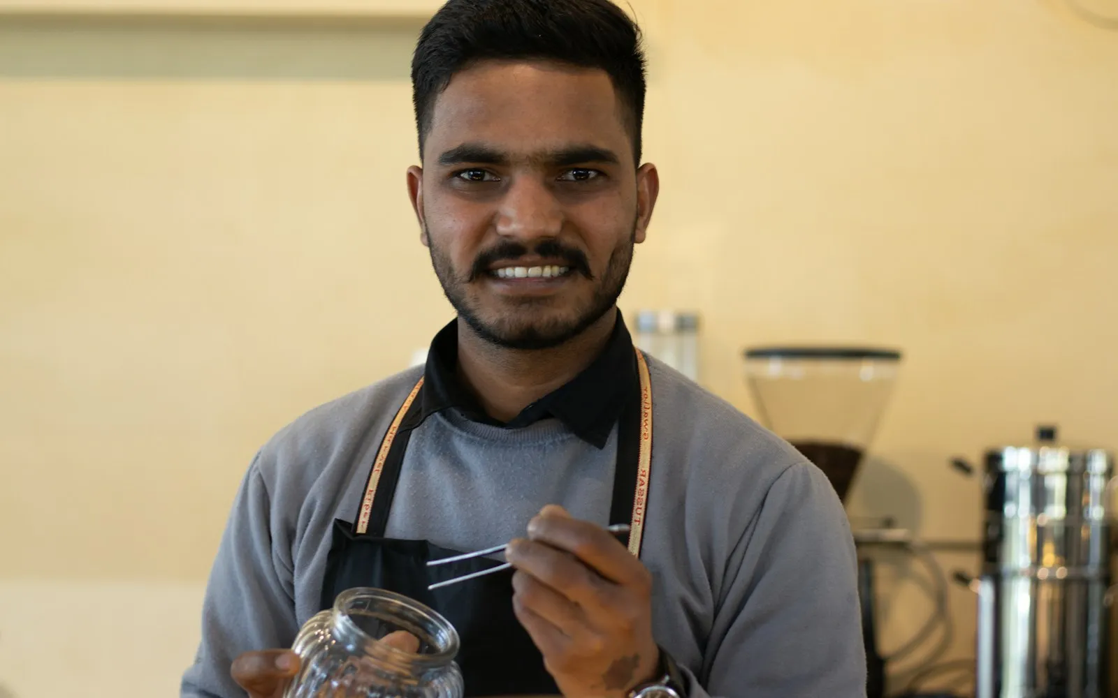Smiling barista in an apron holding a glass jar and tongs in front of a coffee grinder