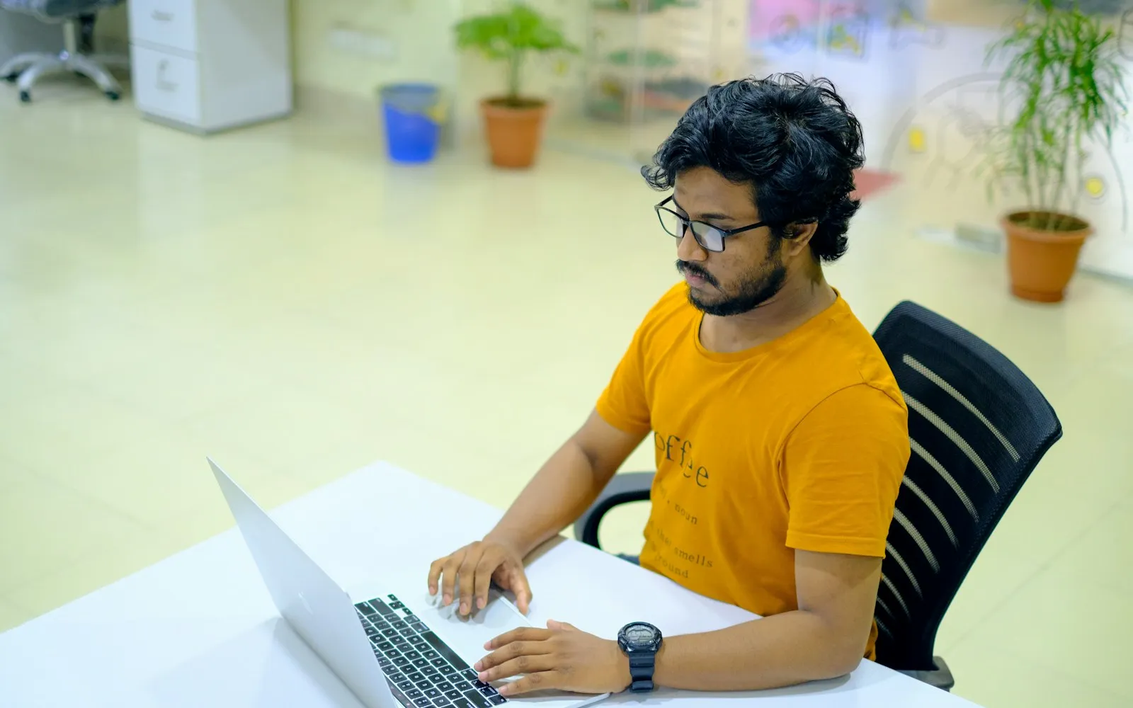 Man in a mustard T-shirt typing on a laptop at a white desk in a bright office