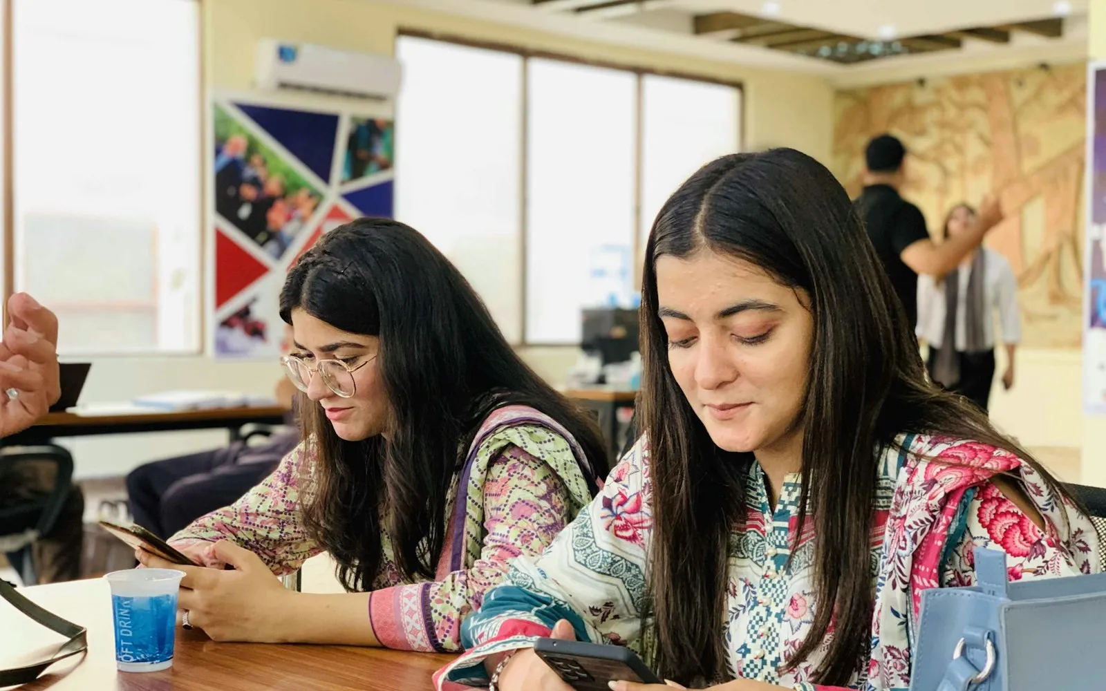 Two women in printed kurtas looking at their smartphones at a table in an office