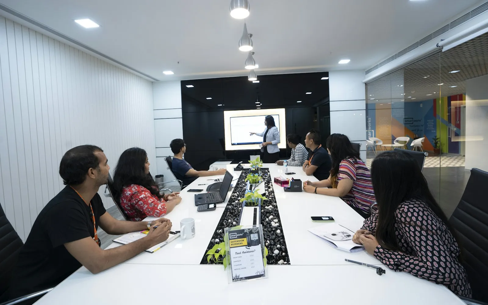 Woman presenting on a screen to colleagues seated around a long white meeting table