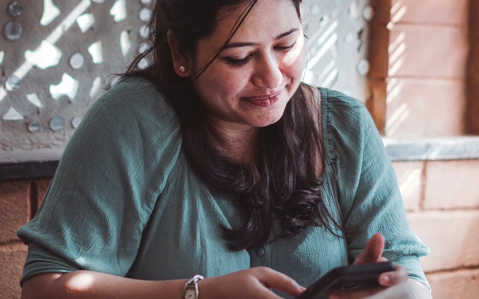 Young Indian woman in a green top smiling at her smartphone at a cafe table with a cup of tea, in warm afternoon light