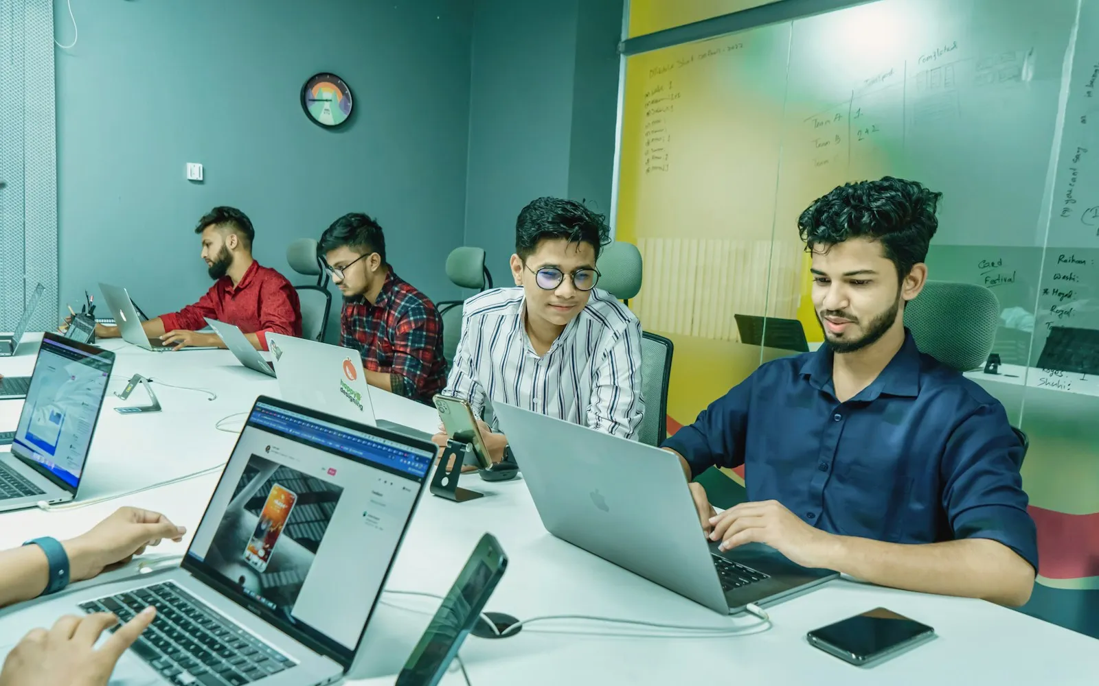 Young team working on laptops around a long conference table with a whiteboard planning wall