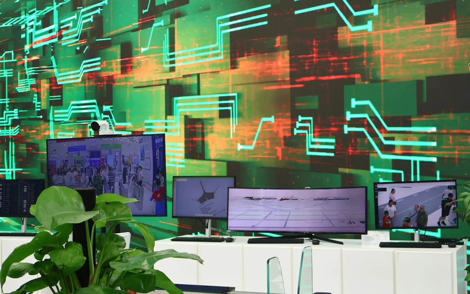 Tech exhibition with monitors showing camera feeds and 3D maps in front of a green circuit-pattern video wall
