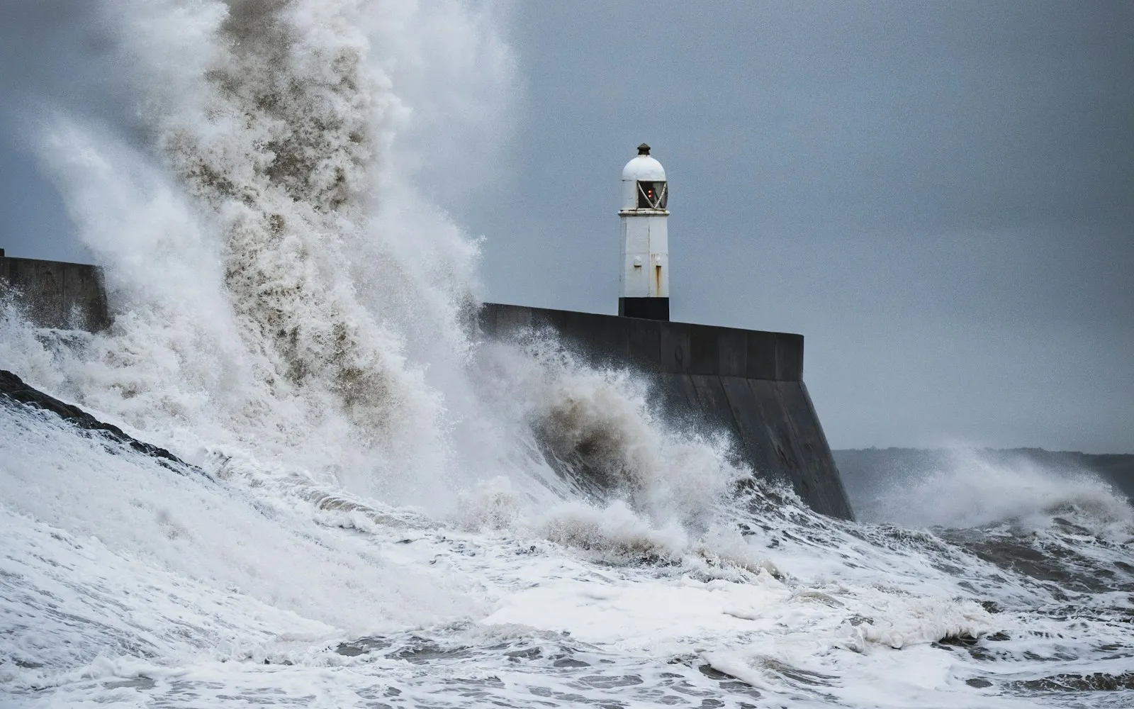 Huge storm waves crashing over a harbour wall and small white lighthouse under grey skies