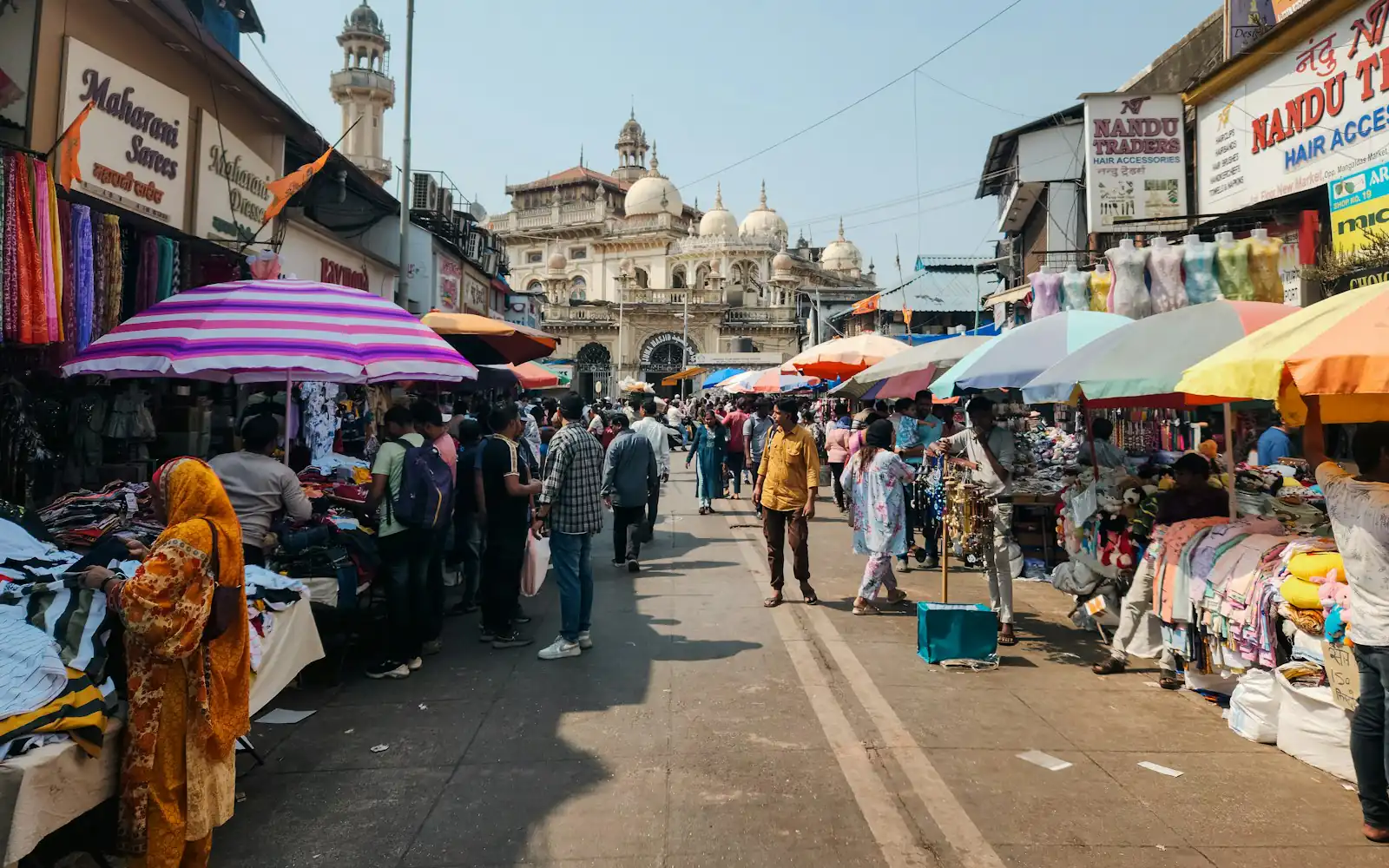 Busy shopping street in Zaveri Bazaar, Mumbai, with clothing stalls under colorful umbrellas, shop signboards and a domed heritage building in the background
