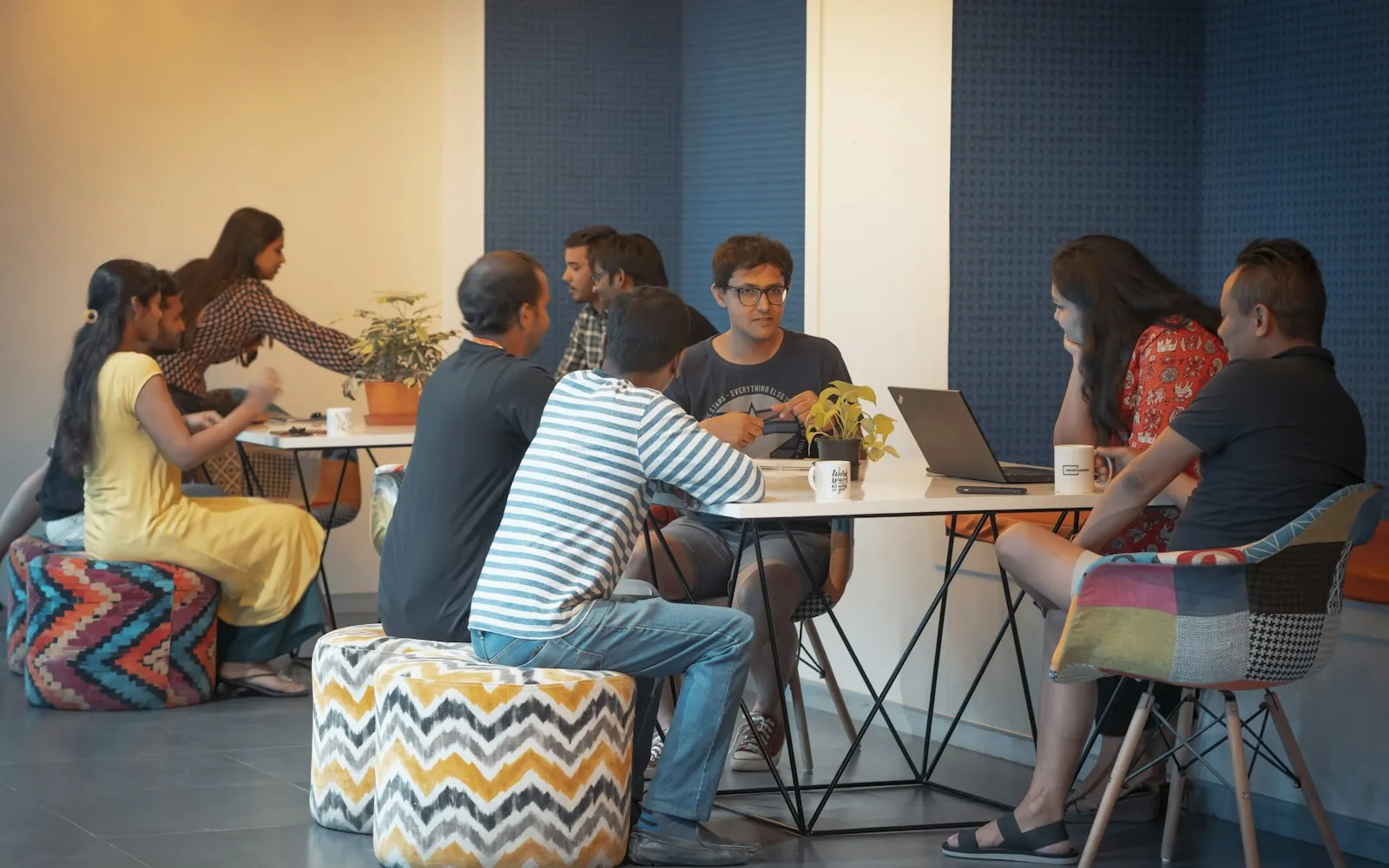 Colleagues in small groups discussing work around tables with patterned poufs in a casual office space