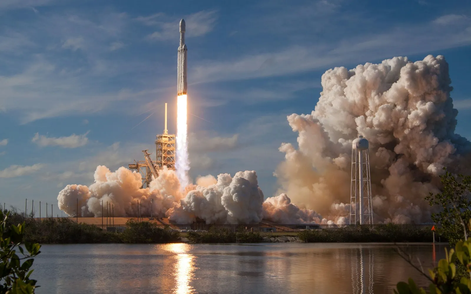 Rocket lifting off from a launch pad in billowing clouds of exhaust, reflected in water