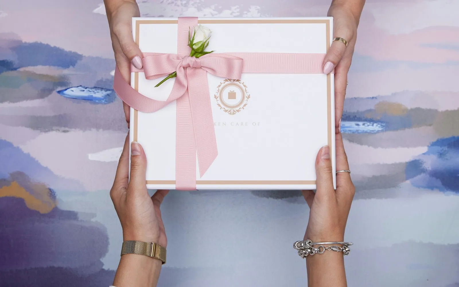 Two pairs of hands exchanging a white gift box tied with a pink ribbon and a white rosebud