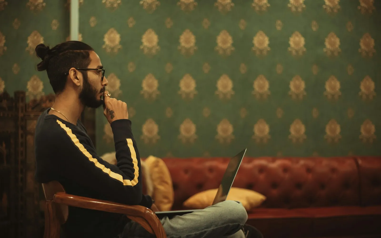 Man with glasses sitting thoughtfully with a laptop on his lap in a room with green damask wallpaper