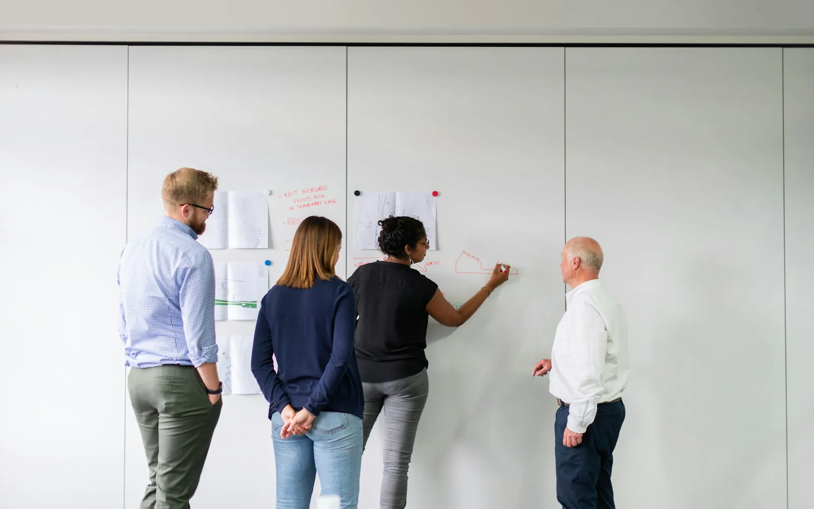 Four colleagues stand at a whiteboard wall in an office while one of them sketches a diagram in red marker beside pinned printouts