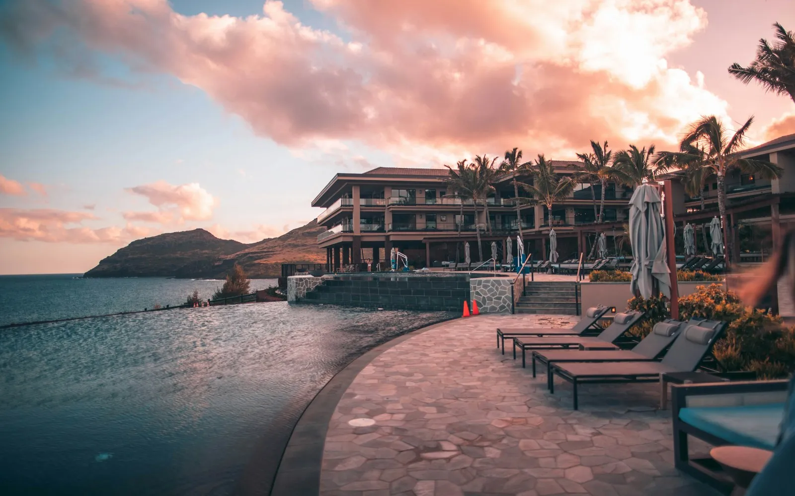 Resort infinity pool and lounge chairs by the ocean at sunset, with palm trees and a hillside
