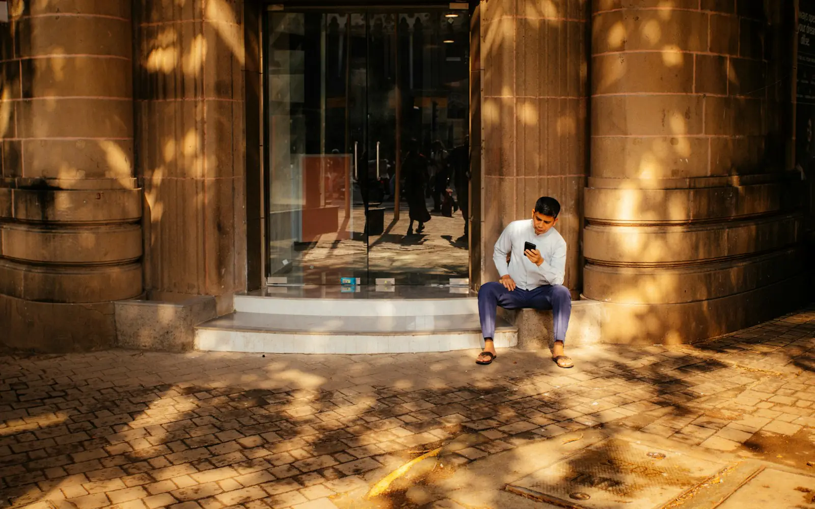 A young man in a white kurta sits on the step of a sunlit stone building in an Indian city, reading something on his phone