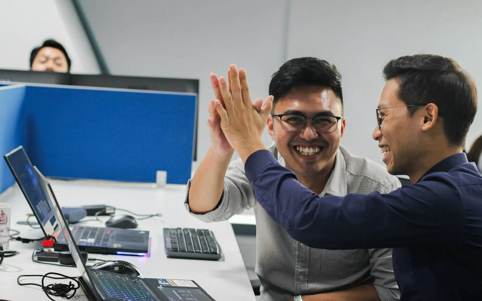 Two smiling colleagues high-fiving at their desks among laptops in an office