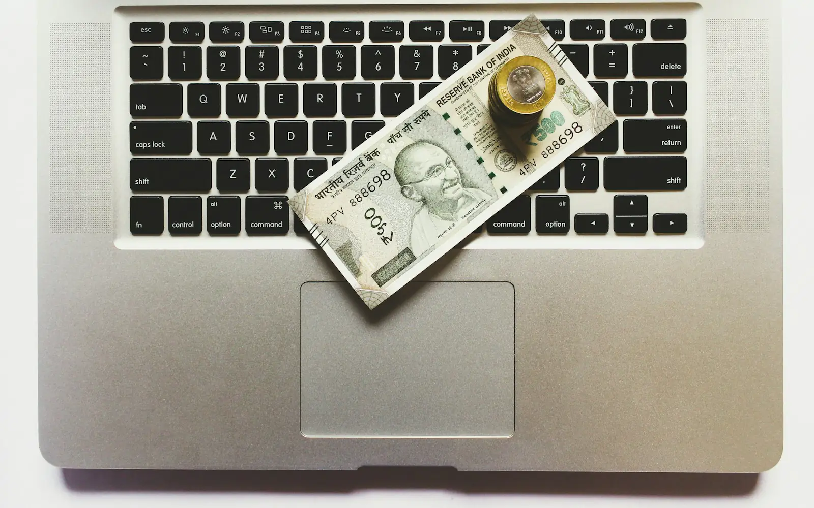 An Indian 500 rupee banknote and a small stack of rupee coins resting on the keyboard of an open silver laptop