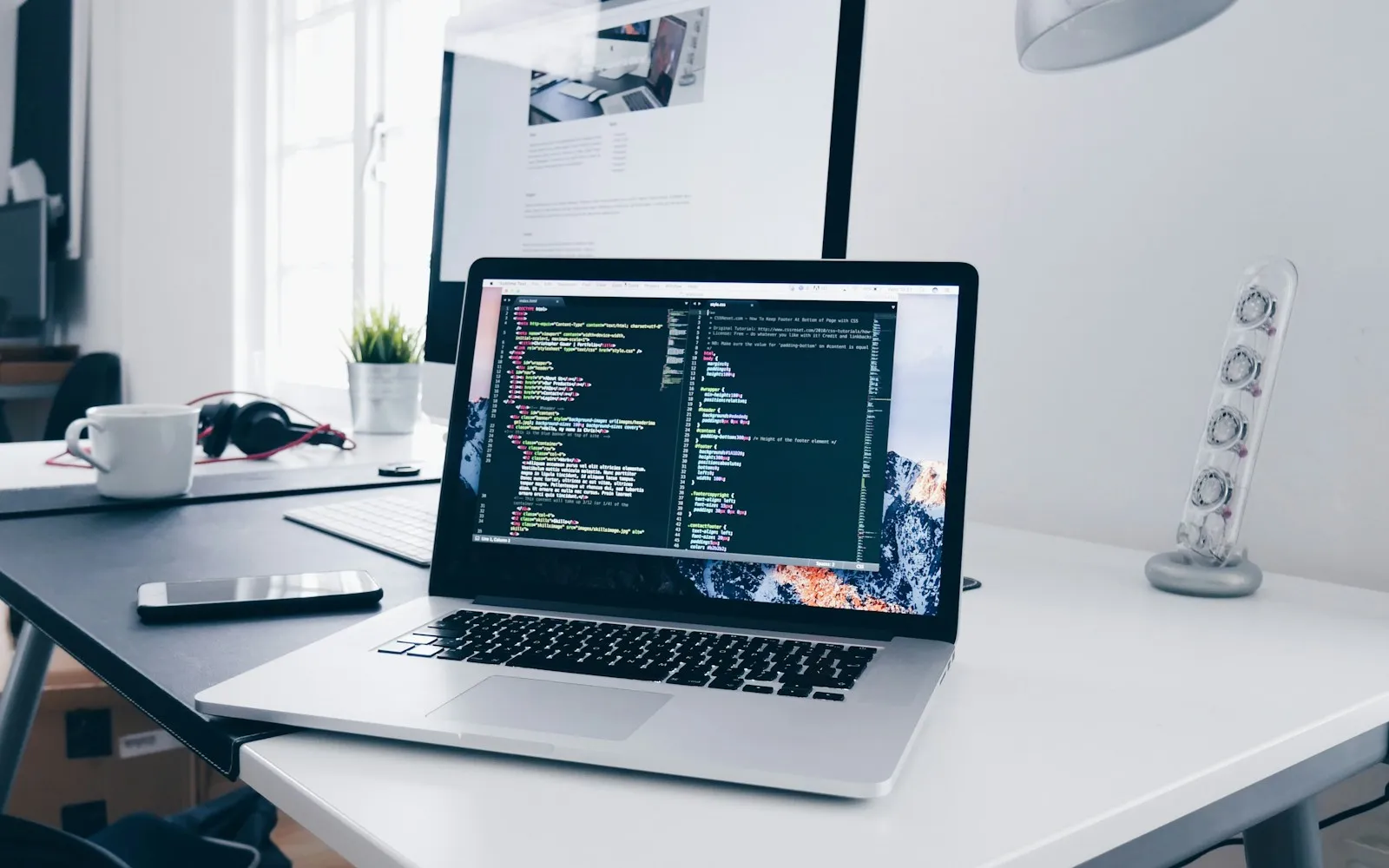 Laptop showing HTML and CSS code on a white desk with a monitor, coffee mug and headphones
