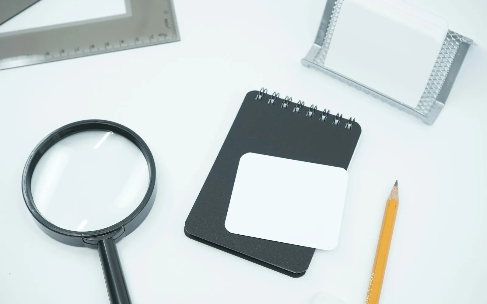 Magnifying glass, black spiral notepad with blank card and a pencil on a white desk
