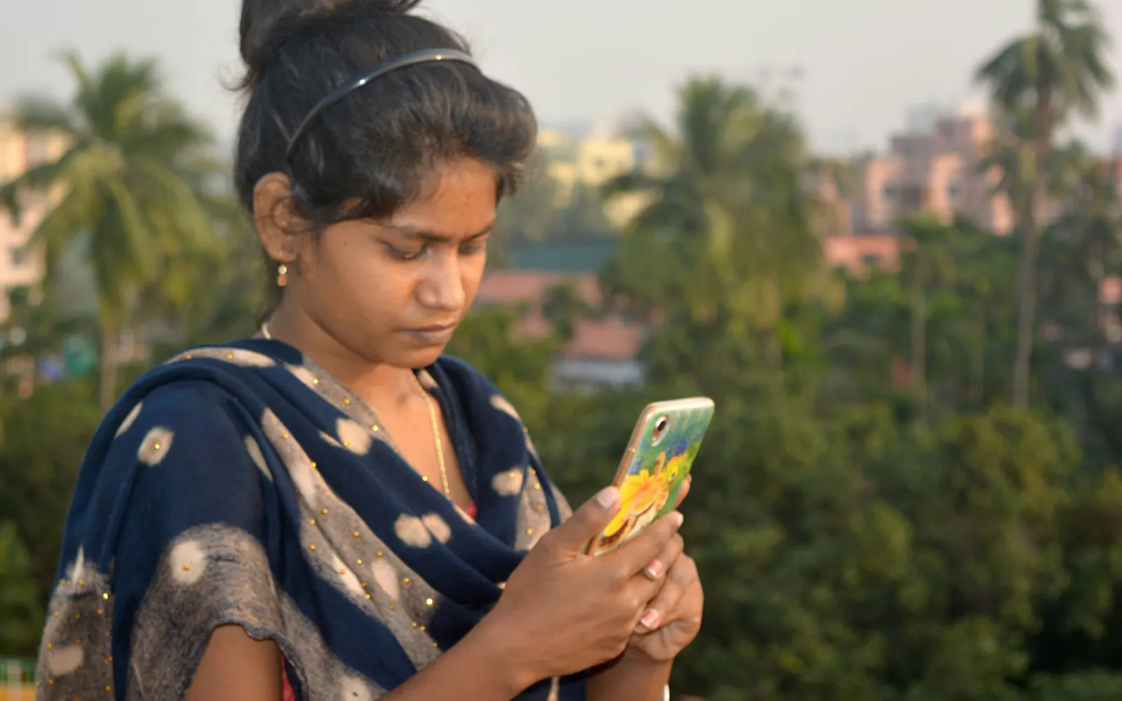 Young Indian woman in a navy dupatta looking at her smartphone on a rooftop, with palm trees and city buildings blurred behind her