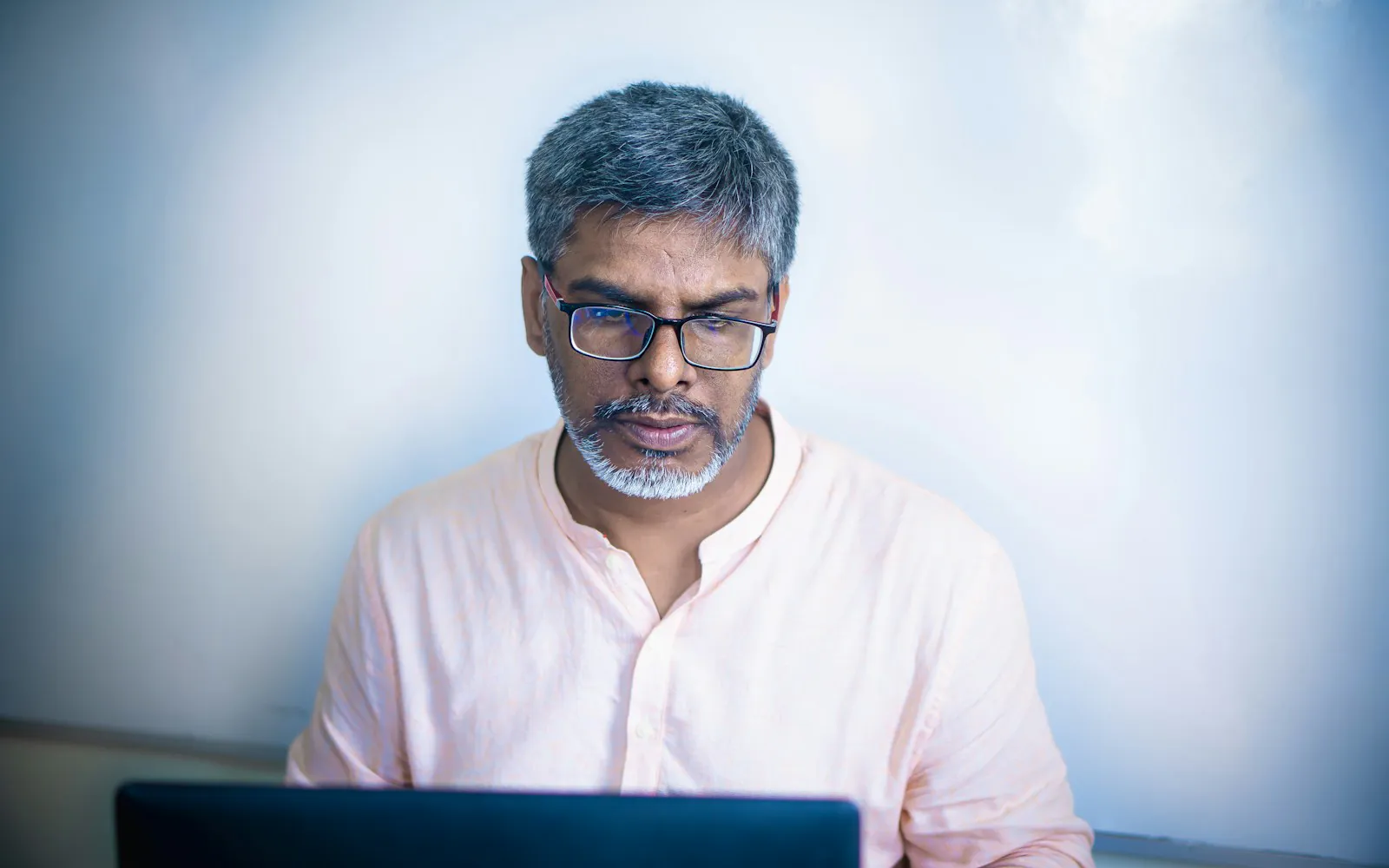 Indian man with grey hair and glasses in a light pink shirt reviewing something on a laptop against a pale blue wall
