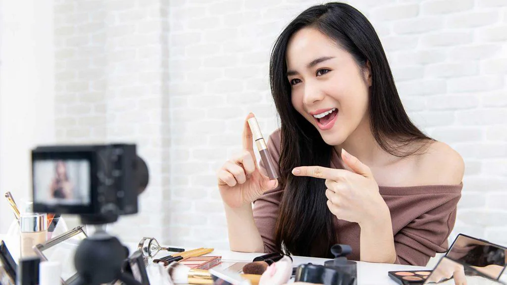 Creator smiling and holding up a lipstick while recording a beauty video on a camera