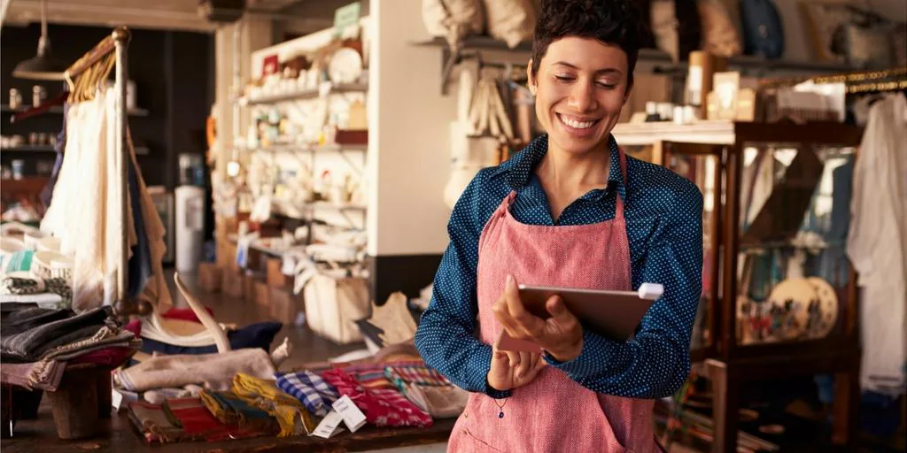 Smiling shop owner in an apron checking orders on a tablet in her store