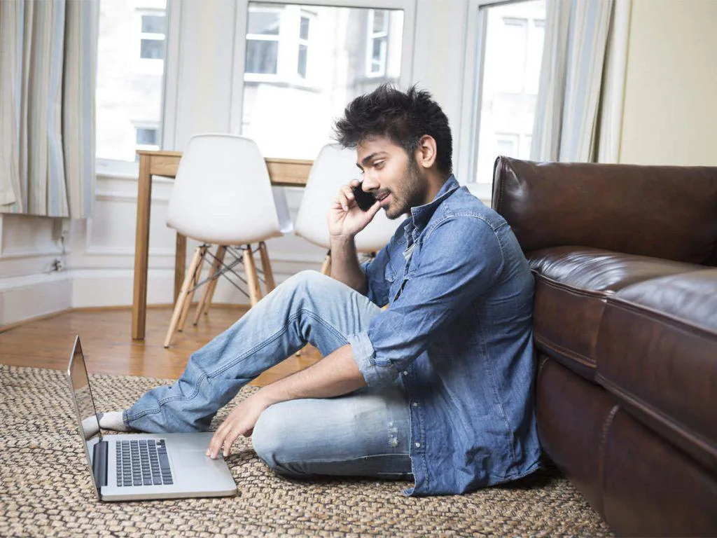 Man working from home on a laptop while taking a phone call, sitting on the floor against a sofa