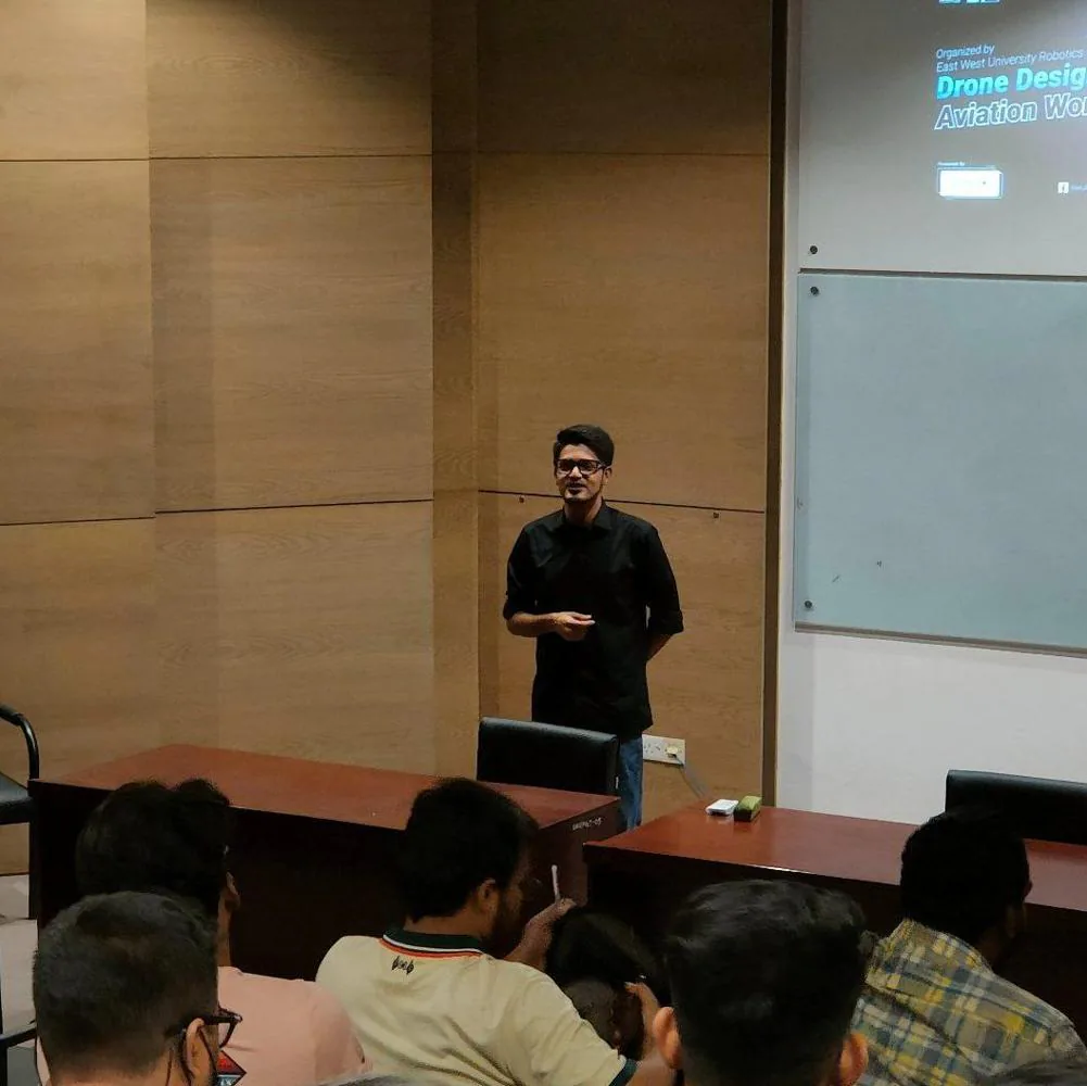 Speaker in a black shirt addressing seated students in a classroom beside a Drone Design workshop slide