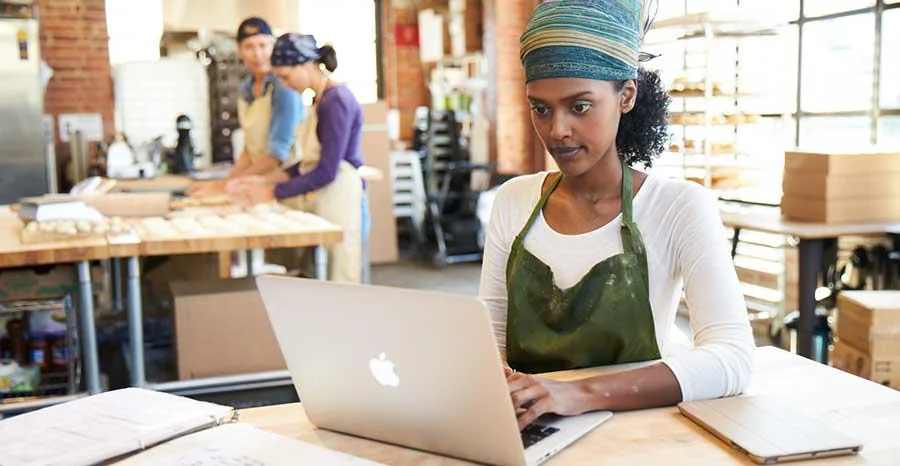 Small business owner working on a laptop in her workshop while colleagues work in the background