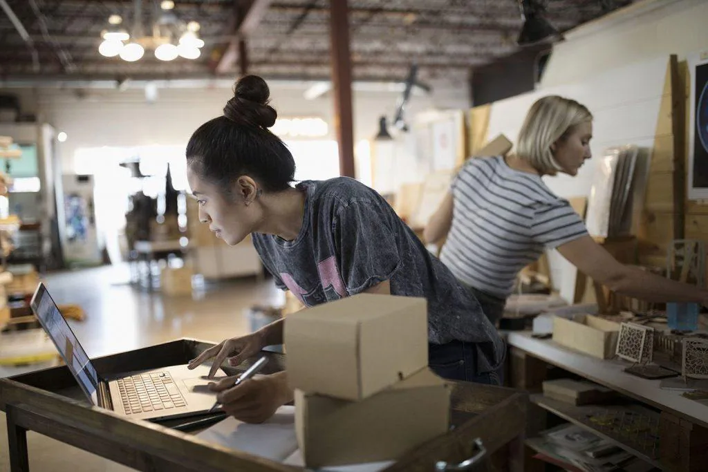 Woman checking orders on a laptop beside shipping boxes while a colleague arranges products in a workshop