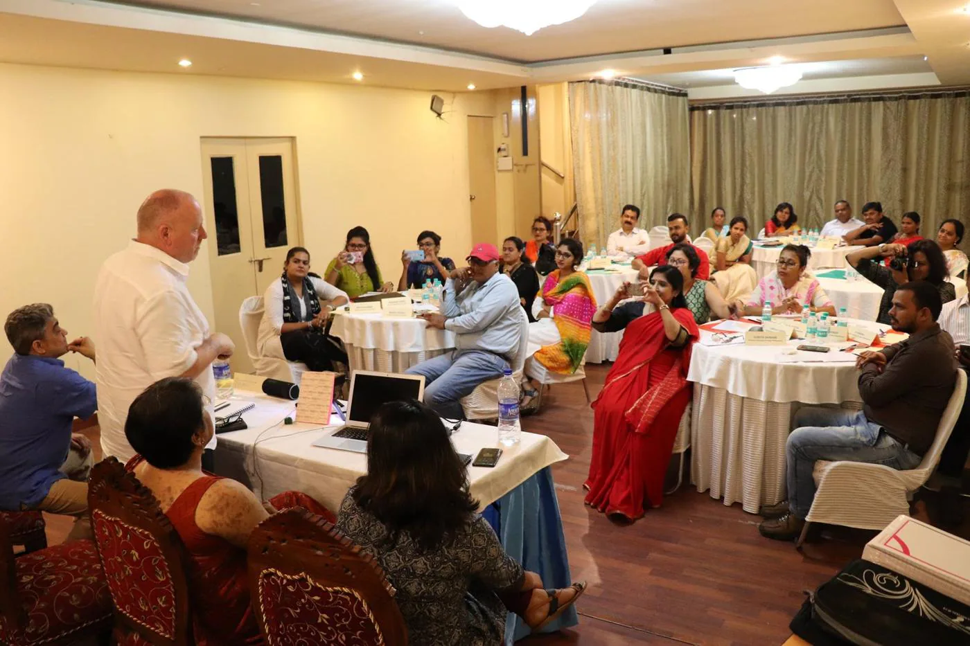 Trainer speaking to a large group of participants seated at round tables in a workshop