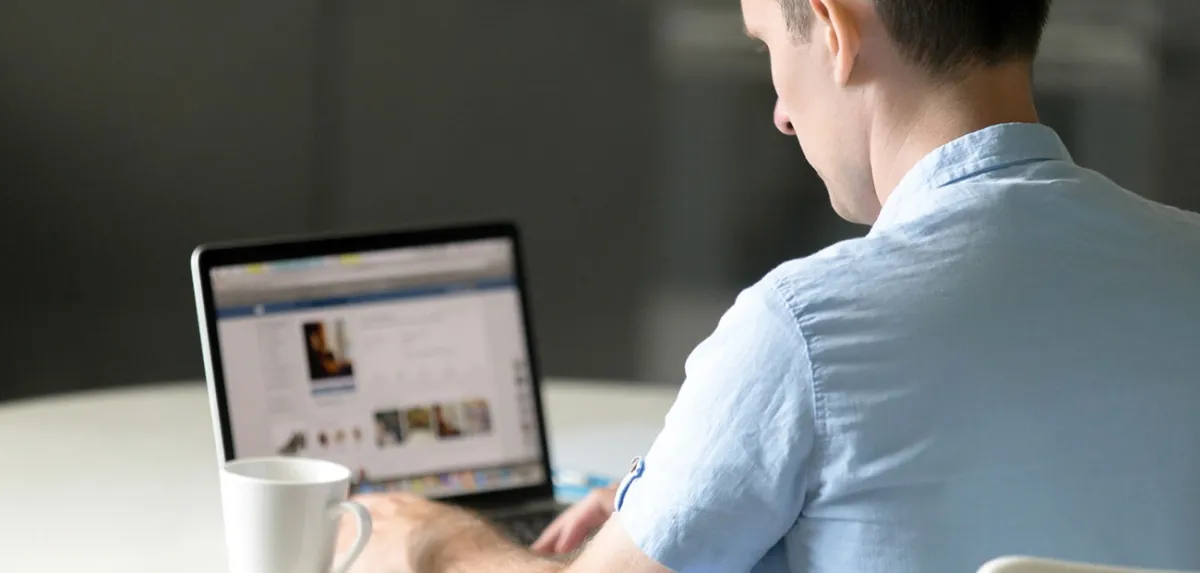 Man in a blue shirt browsing a website on a laptop with a coffee mug on the table