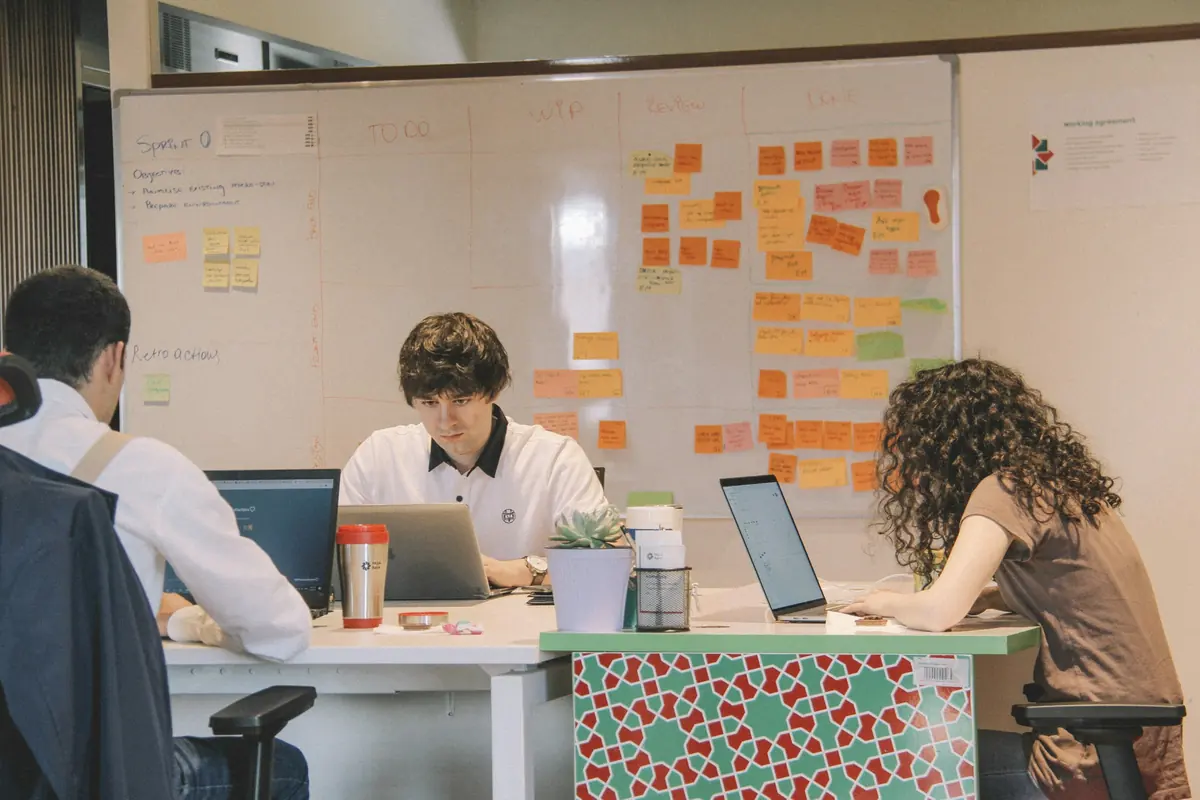 Three team members working on laptops in front of a whiteboard kanban board with To Do, WIP, Review and Done columns
