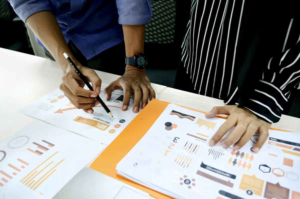 Two people marking up printed charts and infographics with a pencil on a desk