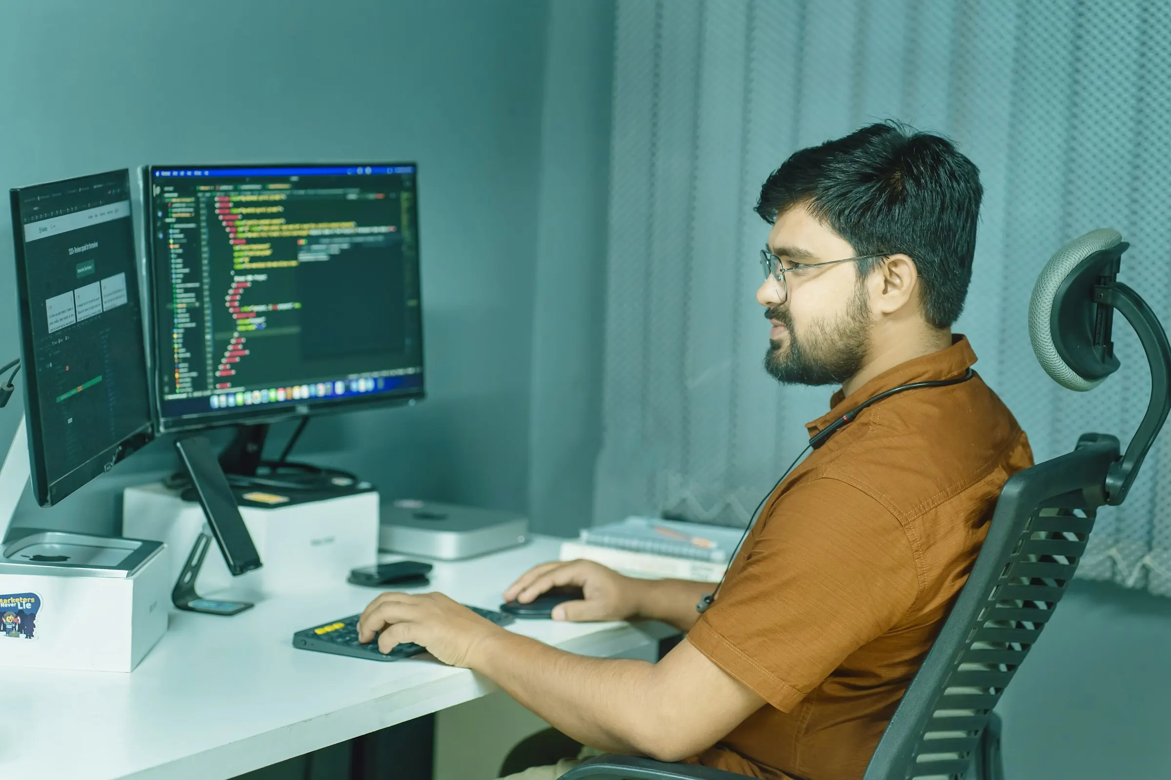 Indian developer in a mustard shirt writing code across two monitors at a white desk