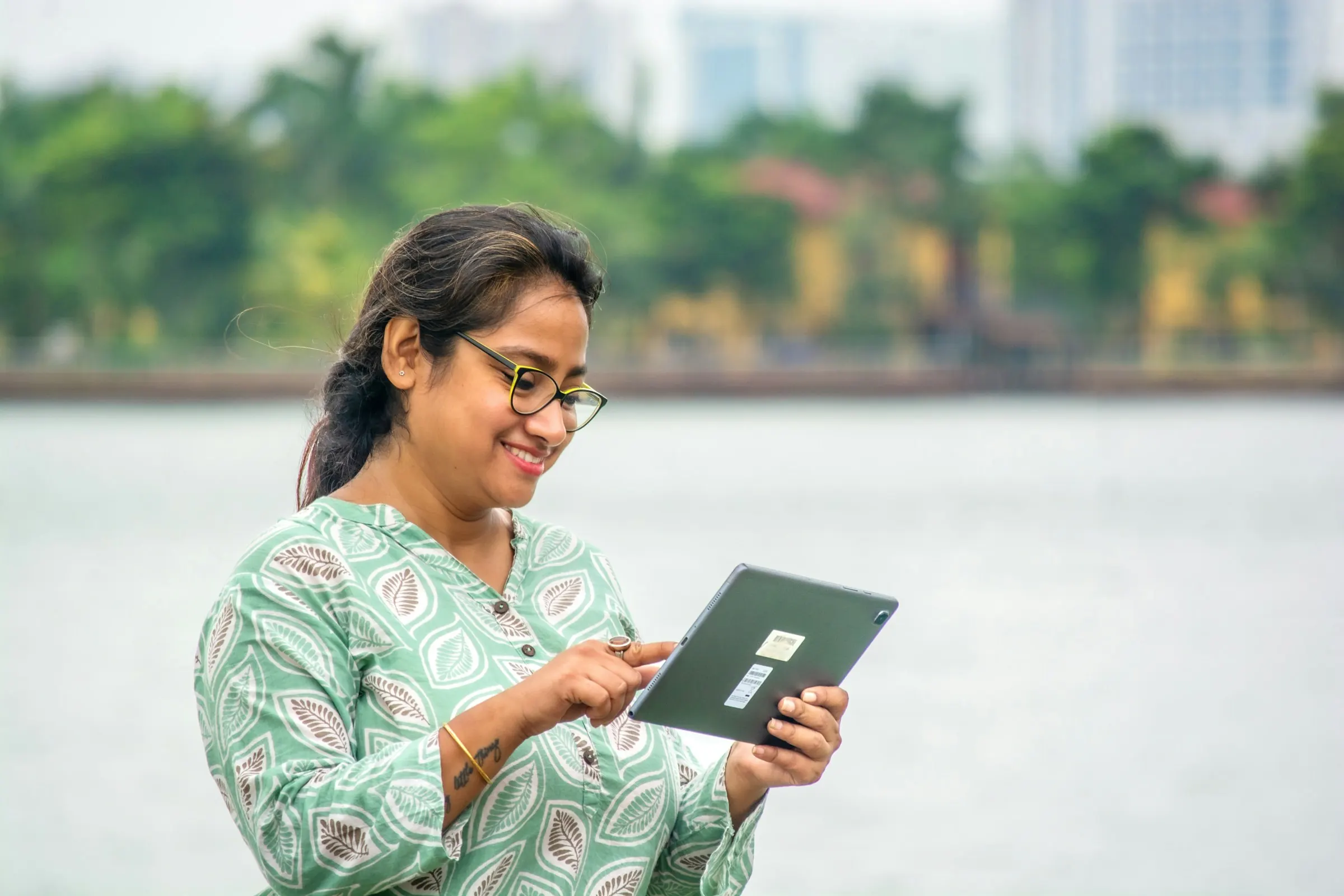 Woman in a kurta smiling as she uses a tablet beside a lake in Kolkata
