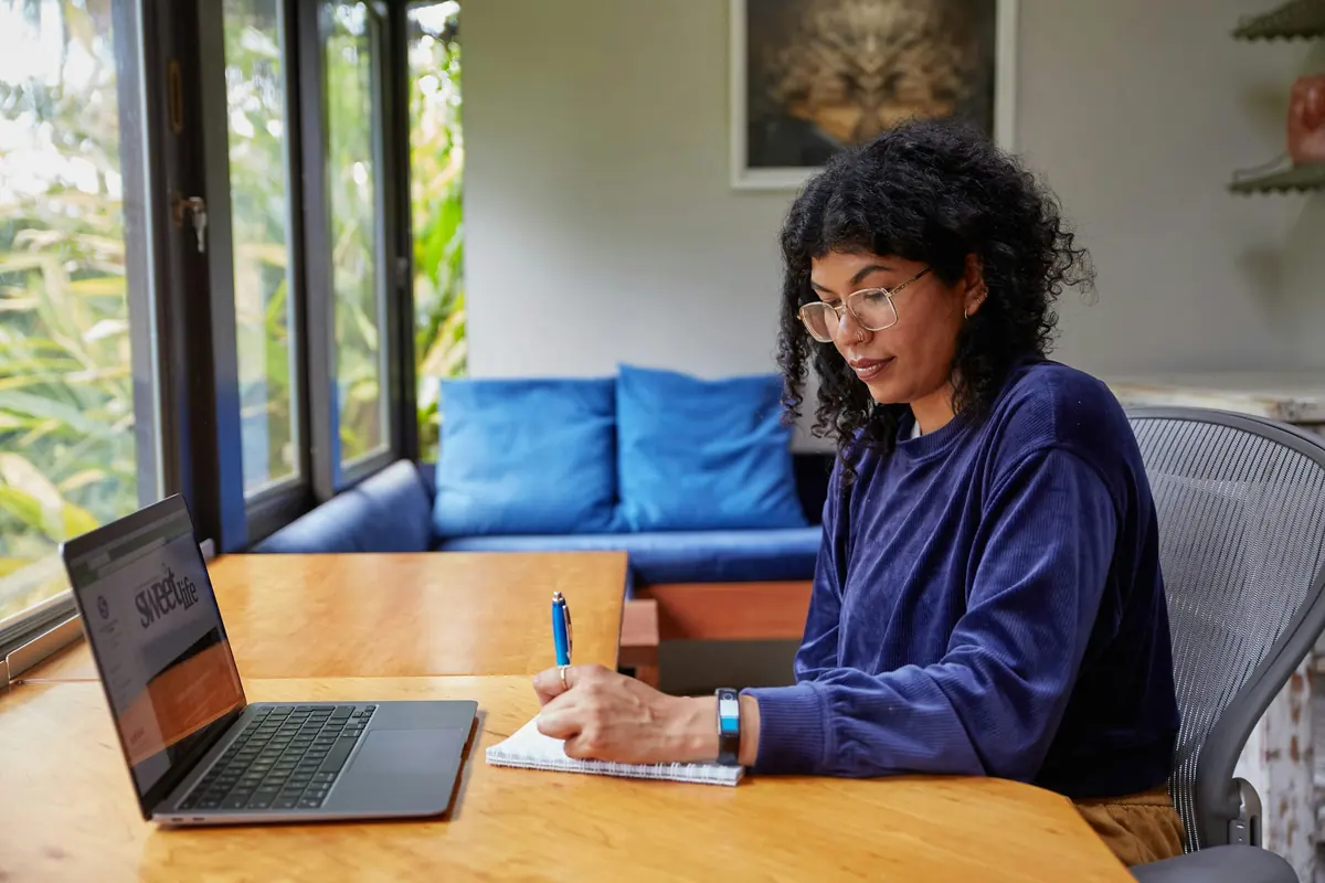 Woman writing notes in a notebook beside an open laptop at a wooden table by a window